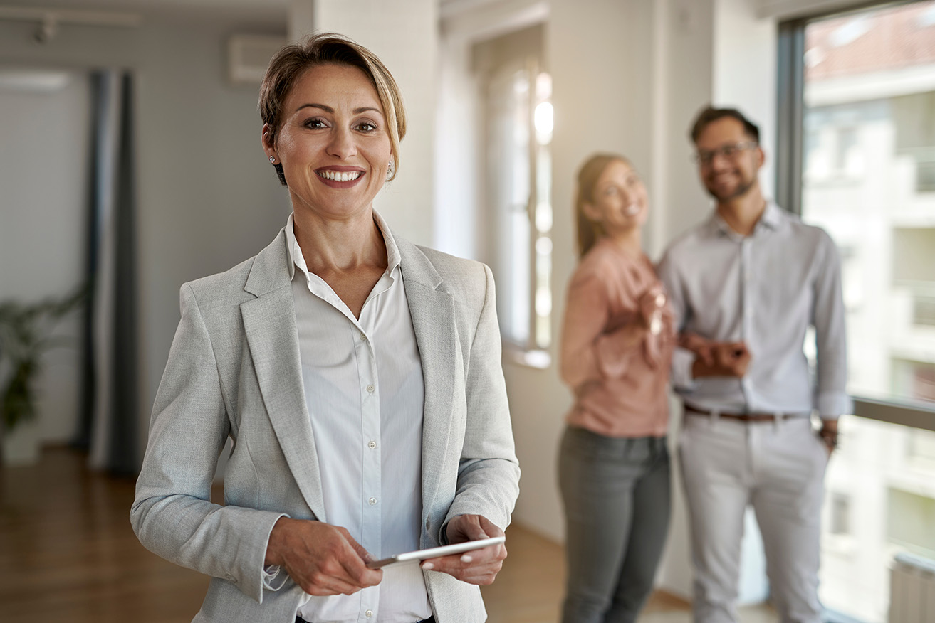 Smiling professional woman in a light gray blazer holding a tablet, with two people discussing in the background.