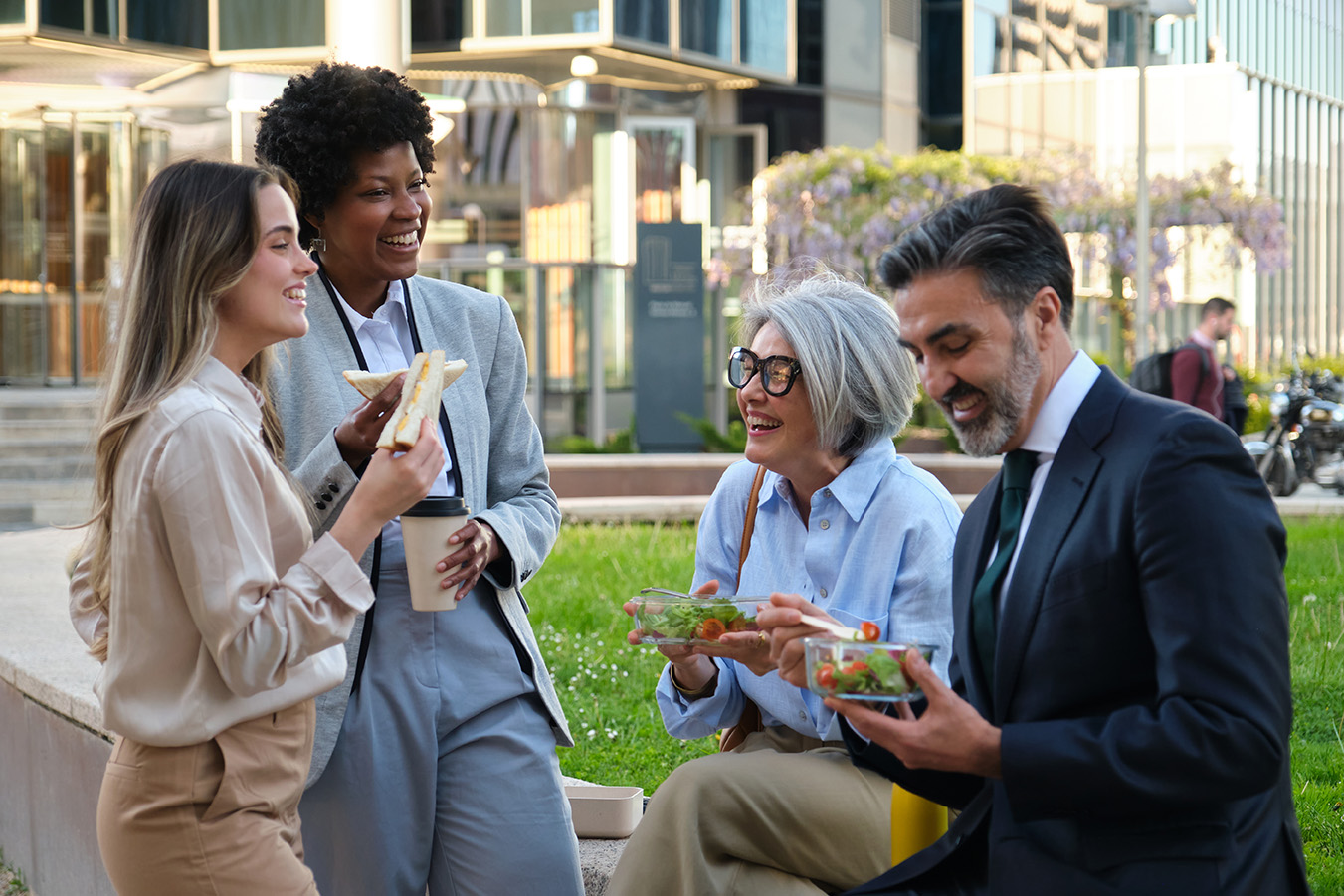 A diverse group of colleagues enjoying lunch together outdoors, smiling and sharing food.