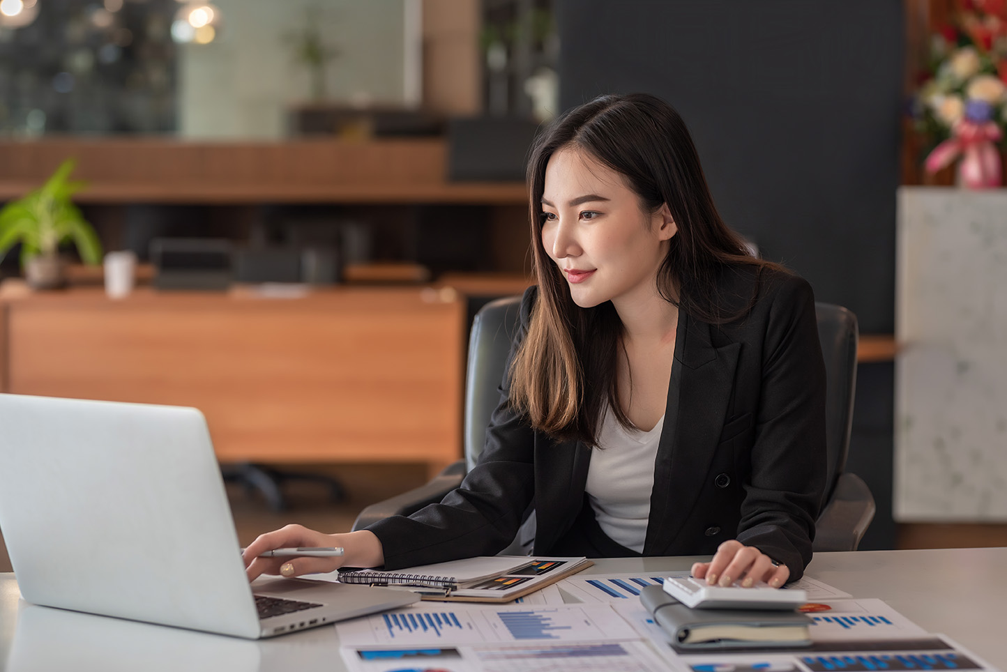 Young businesswoman in a black blazer working on a laptop with financial charts and reports on her desk.