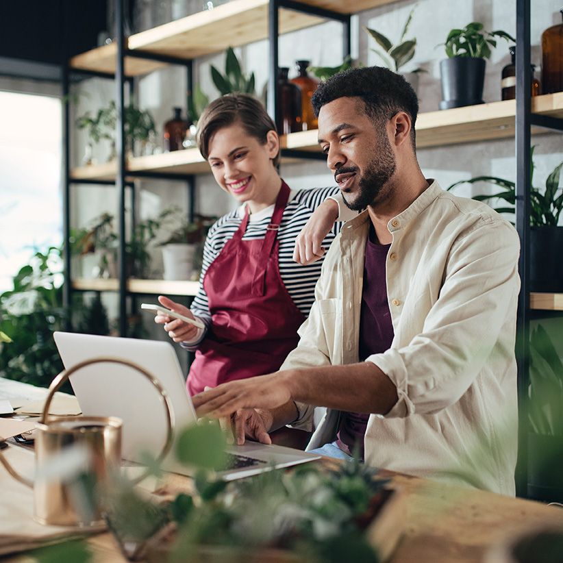 Two small business owners working together on a laptop in a modern plant-filled workspace, collaborating on tasks.